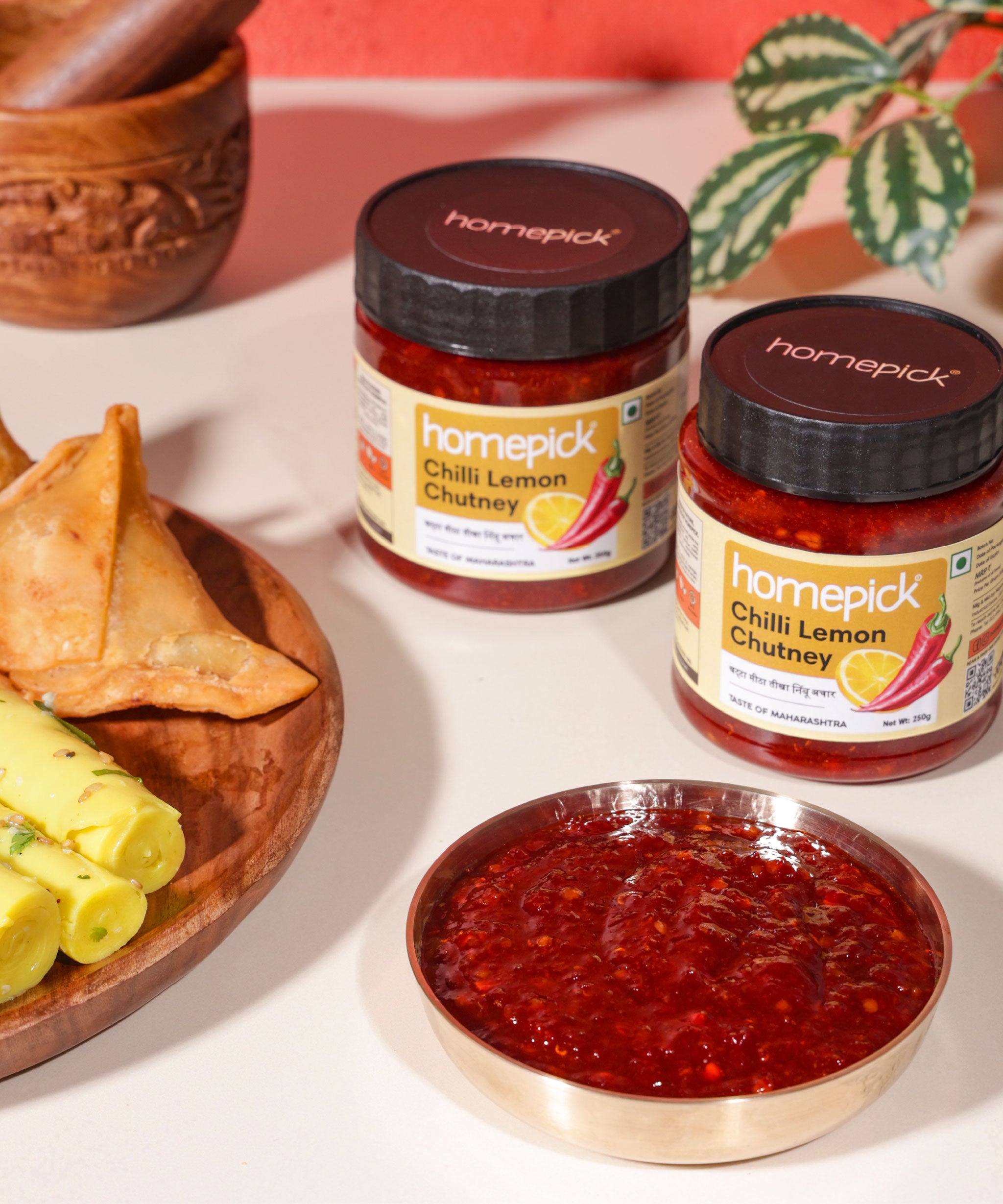 Two jars of homepick Chilli Lemon Chutney with a bowl of chutney and samosas on a white surface.
