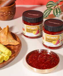 Two jars of homepick Chilli Lemon Chutney with a bowl of chutney and samosas on a white surface.